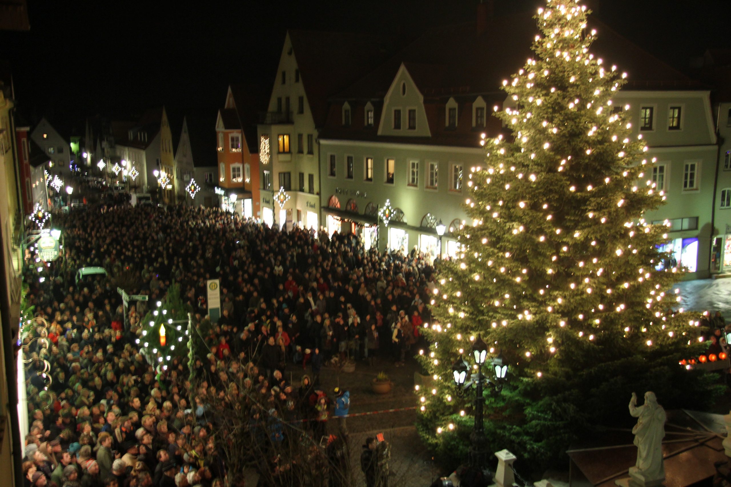 Bild Blick auf Marktplatz in der Nacht, im Vordergrund beleuchteter Weihnachtsbaum mit Menschenmenge.